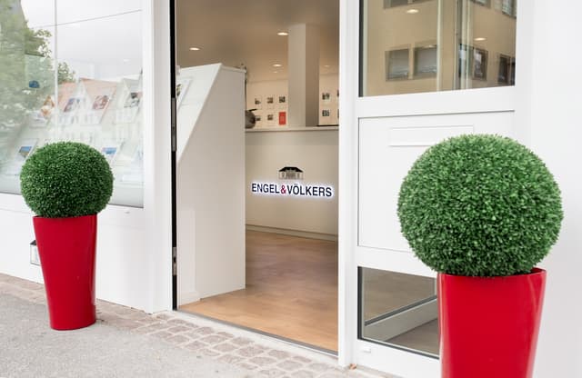 Engel & Völkers office entrance with two round green bushes in red pots flanking the open doorway. The logo is visible inside.