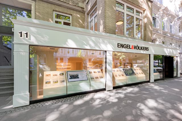 Exterior view of Engel & Völkers real estate office with large windows displaying property listings. The building is brick with white trim.