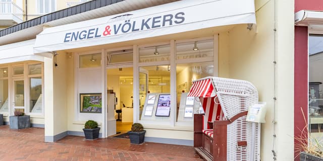 Engel & Völkers real estate office exterior with a white awning, display windows, and a red and white striped beach chair.