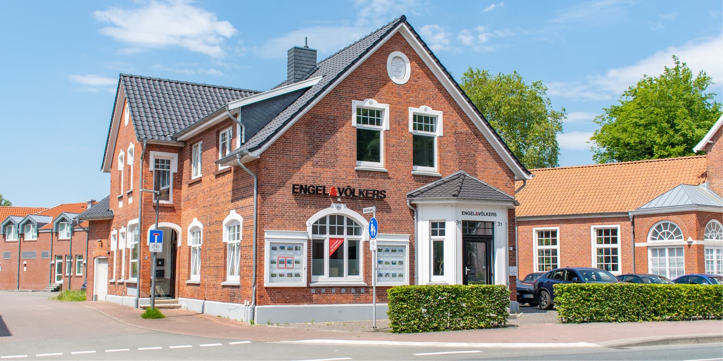 Engel & Völkers real estate office in a red brick building with a dark gray roof on a sunny day. Cars are parked in front.