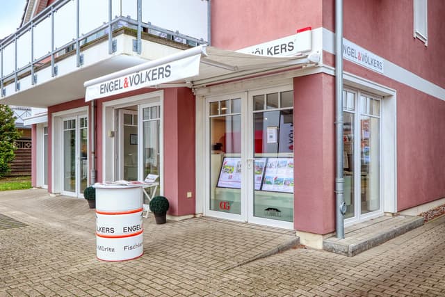 Exterior of Engel & Völkers real estate office, pink building with white trim, brochures in windows, barrel table.