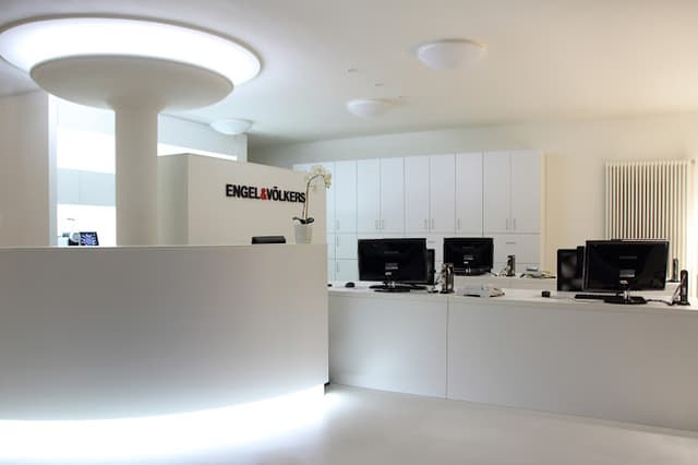 Engel & Völkers Büro-Interieur. Weißer Empfangstresen mit Logo, Computern und Schränken. Moderne Beleuchtung.