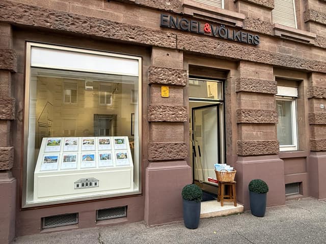 Exterior of Engel & Völkers real estate office with stone facade. Window display shows property listings. Open door with brochures.