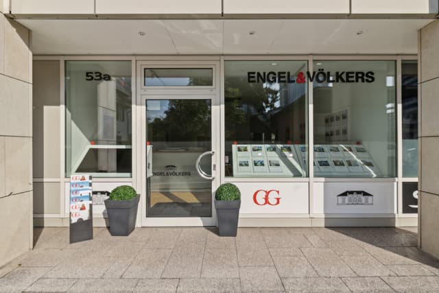 Engel & Völkers real estate office storefront with glass windows and door. "53a" address, logo, and property listings are visible. Two potted plants flank the entrance.
