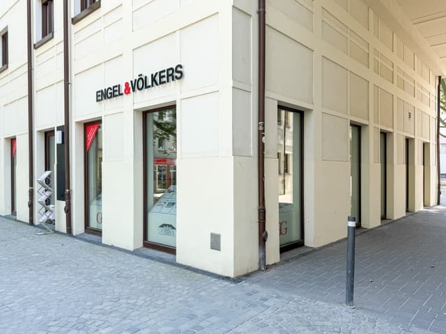 Engel & Völkers real estate office on a corner. The building is white with dark-framed windows. A gray cobblestone street is in front.