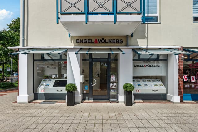 Engel & Völkers real estate office storefront with glass windows and door. Two potted plants flank the entrance.