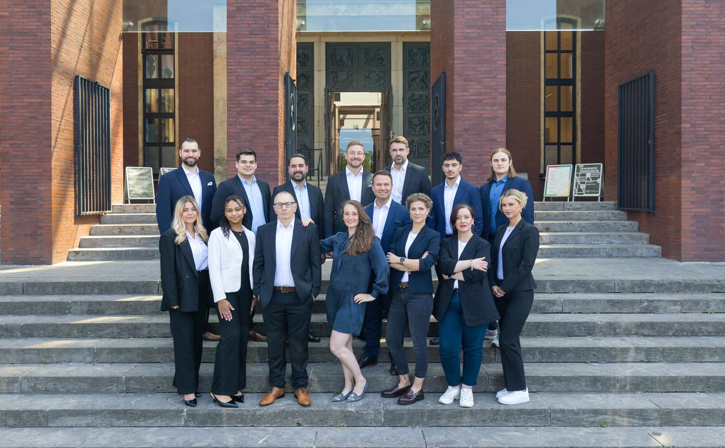 A group of 15 real estate agents stand on stone steps in front of a brick building with large doors. They wear business attire.