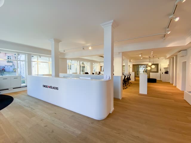 Bright real estate office with wood floors and white walls. A curved white reception desk with the Engel & Völkers logo is in the foreground.