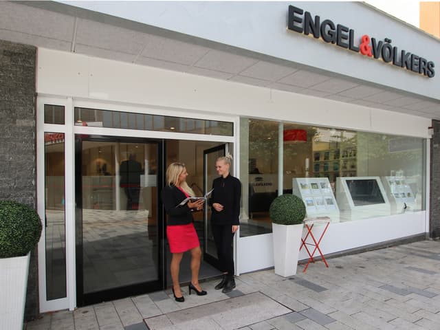Two women stand outside an Engel & Völkers real estate office. One in a red dress holds a brochure, the other in black.