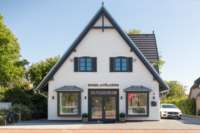 Engel & Völkers Immobilienbüro. Weißes Gebäude mit schwarzen Akzenten, Fenstern und Dach. Auto rechts geparkt.