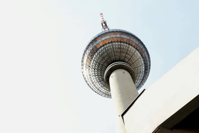 Looking up at the Berlin TV Tower, a tall, gray structure with a red and white antenna against a pale blue sky.