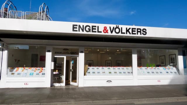 Engel & Völkers real estate office with a white facade and large windows displaying property listings. Open door on the left.