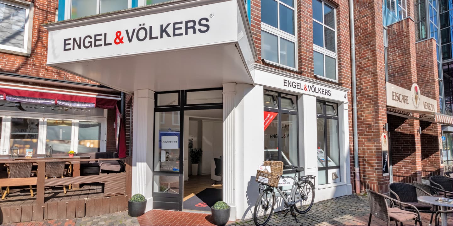 Engel & Völkers real estate office exterior. A bicycle with baskets sits outside the white building with black framed windows. Brick buildings surround.