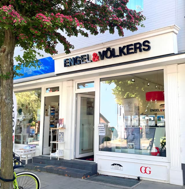 Engel & Völkers Immobilienbüro mit weißer Verkleidung und schwarzer Beschriftung. Links steht ein Baum mit rosa Blüten.