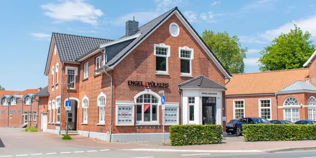 Engel & Völkers real estate office in a red brick building with a dark gray roof on a sunny day. Cars are parked in front.