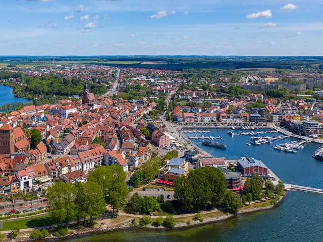 Aerial view of Waren, Germany, featuring red-roofed buildings, a harbor with boats, and a blue sky with scattered clouds.
