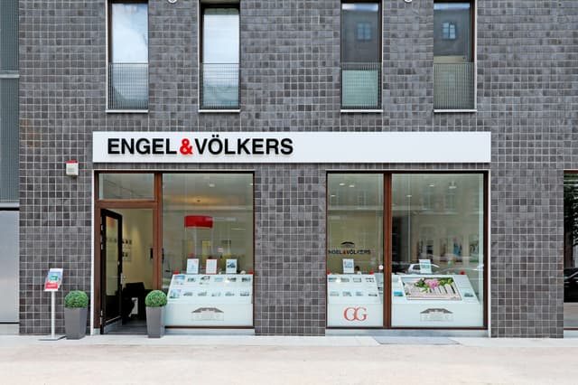 Engel & Völkers real estate office front with a gray tiled facade, large windows displaying property listings, and an open doorway.