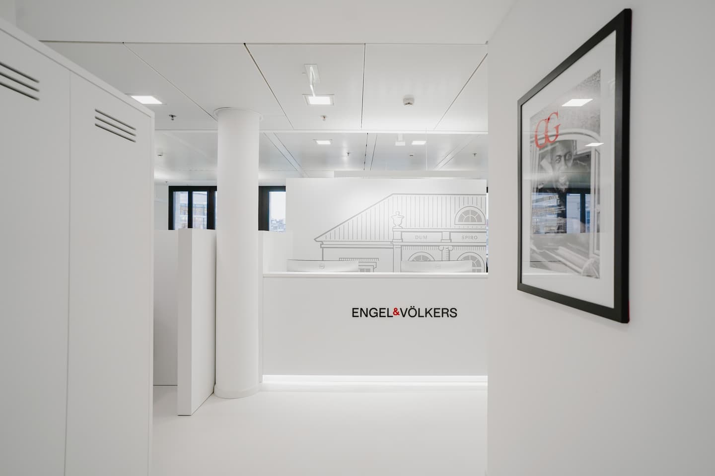 Bright, modern Engel & Völkers office reception area. White desk with logo, monitors, and orchid plant. Building sketch on wall.