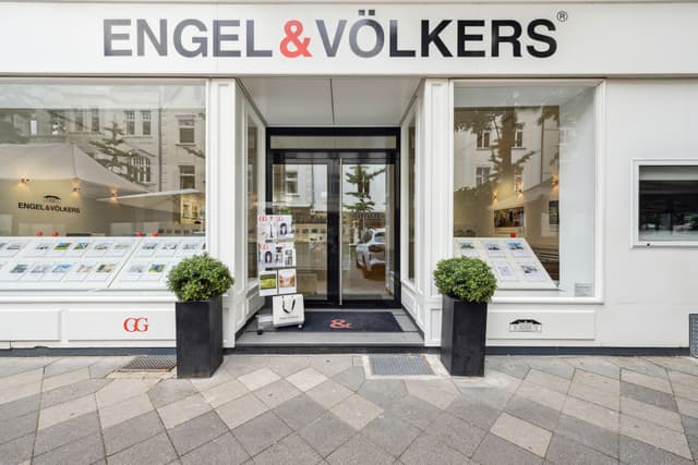 Engel & Völkers real estate office with a white facade, glass windows displaying property listings, and black planters with green bushes.