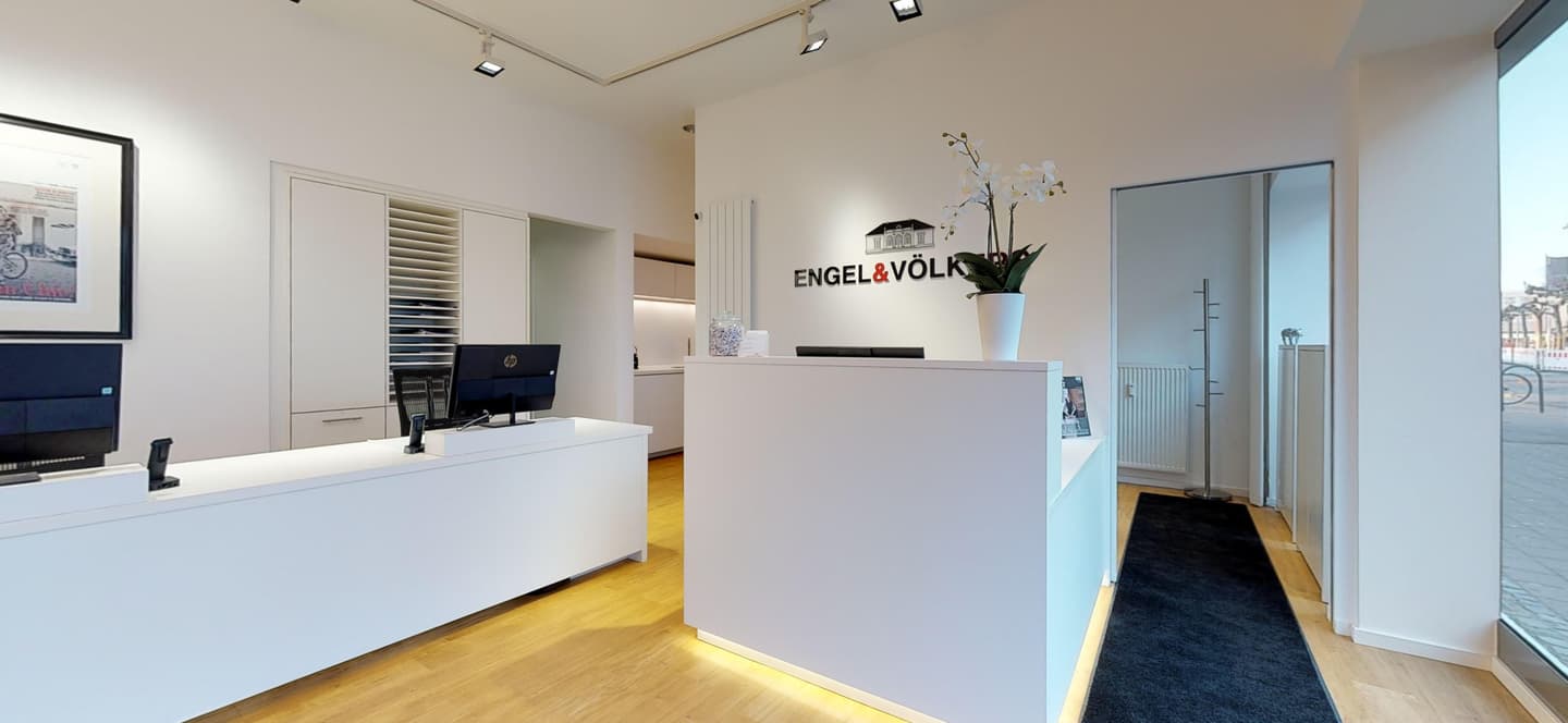 Engel & Völkers real estate office interior. White reception desk with orchid, logo on wall, wood floors, and a view to the outside.