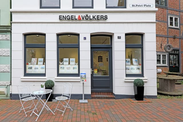 Exterior of Engel & Völkers real estate office with white facade, dark gray door and windows. A table and chairs sit on a brick walkway.