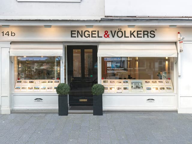 Engel & Völkers real estate office storefront. White building with black door, awnings, and property listings in the windows.