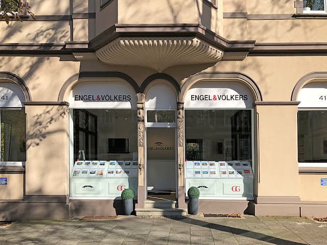 Engel & Völkers real estate office with arched windows displaying property listings. Beige building with potted plants.