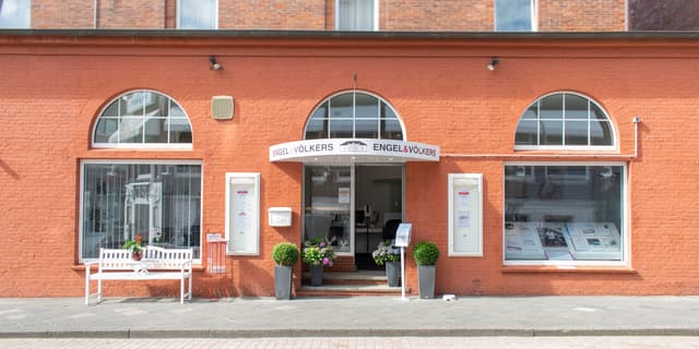 Engel & Völkers real estate office with a red brick facade and arched windows. A white bench sits outside.