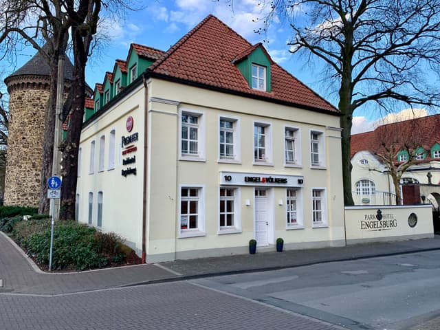 Exterior view of a cream-colored, two-story building with a red tile roof and green dormers, Engel & Volkers signage, and a stone tower nearby.