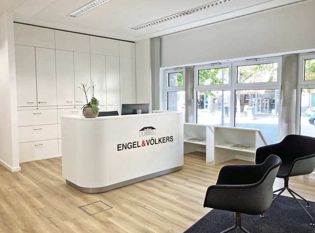 Bright real estate office with white reception desk, Engel & Völkers logo, black chairs, and light wood floors. Large windows show street view.