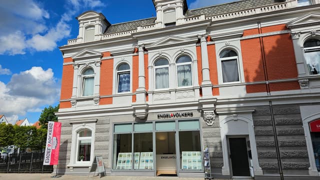 Außenansicht des Engel & Völkers Immobilienbüros, ein rotes Backsteingebäude mit weißen Elementen unter blauem Himmel mit Wolken.