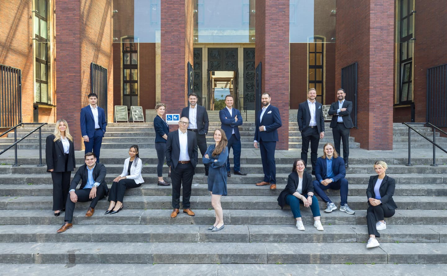 A group of 14 real estate agents stand on stone steps in front of a building with large, ornate bronze doors. Most wear business attire.