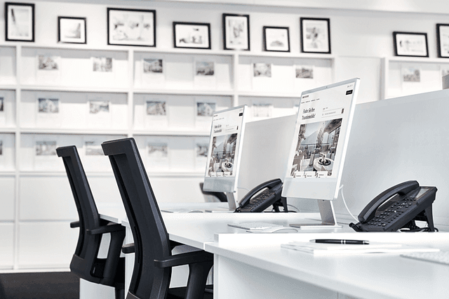 A bright real estate office with white desks, black chairs, computers, and phones. A wall of framed property photos is in the background.