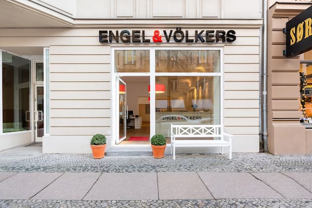 Engel & Völkers real estate office exterior. White bench and potted plants flank the glass storefront with black lettering.