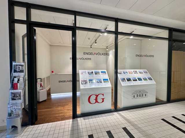 Engel & Völkers real estate office with black framed glass windows and door. White displays show property listings. Tiled floor.