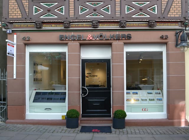 Engel & Völkers real estate office with a black door, flanked by windows displaying property listings. The building has a brown facade and decorative carvings.