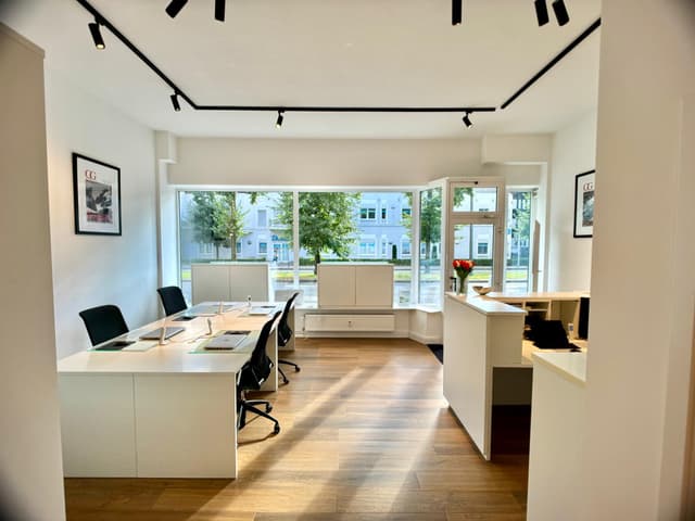 Bright real estate office with white walls, wood floors, and large windows. Desks with laptops and black chairs face the window. A reception desk is to the right.