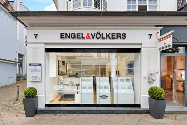 Engel & Völkers real estate office storefront with large glass windows and white trim. Planters flank the entrance.
