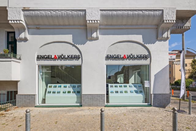 Engel & Völkers real estate office with two arched windows displaying property listings. White building with decorative molding.