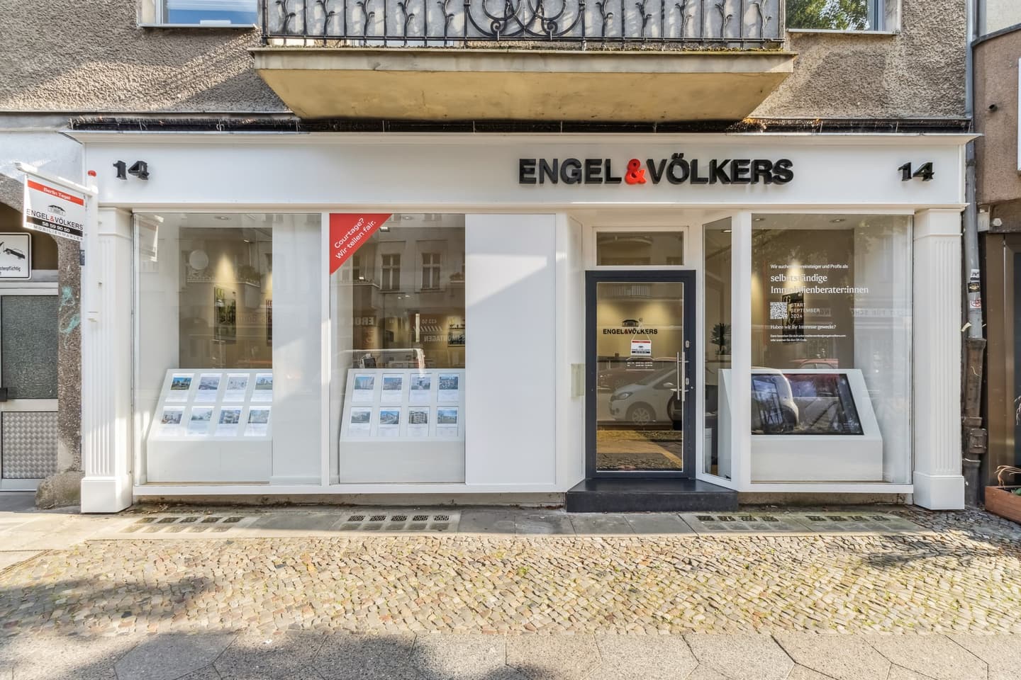 Engel & Völkers Immobilienbüro mit weißer Verkleidung und großen Fenstern, in denen Immobilienangebote ausgestellt sind. Kopfsteinpflaster-Gehweg davor.