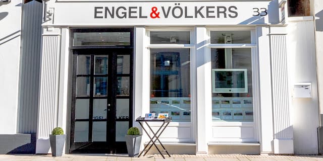 Engel & Völkers Immobilienbüro mit schwarzer Tür, weißen Zierleisten und Immobilienangeboten im Fenster.