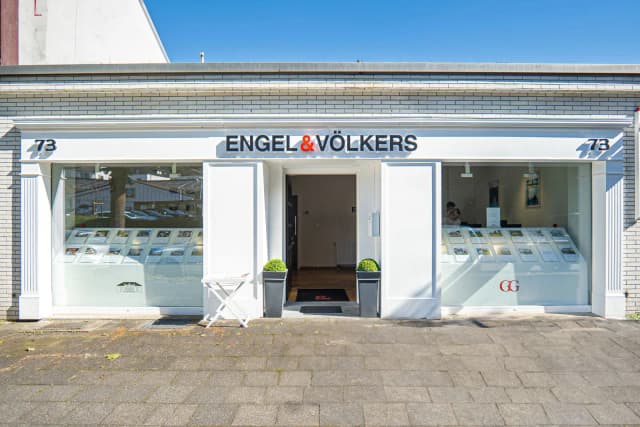 Engel & Völkers real estate office with white brick facade, large windows displaying property listings, and an open doorway.