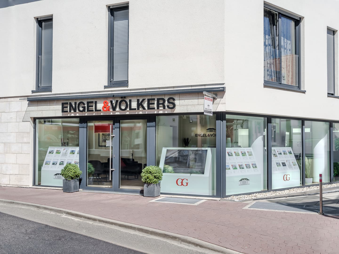 Exterior of Engel & Völkers real estate office with black framed windows and doors. A display of property listings is visible.