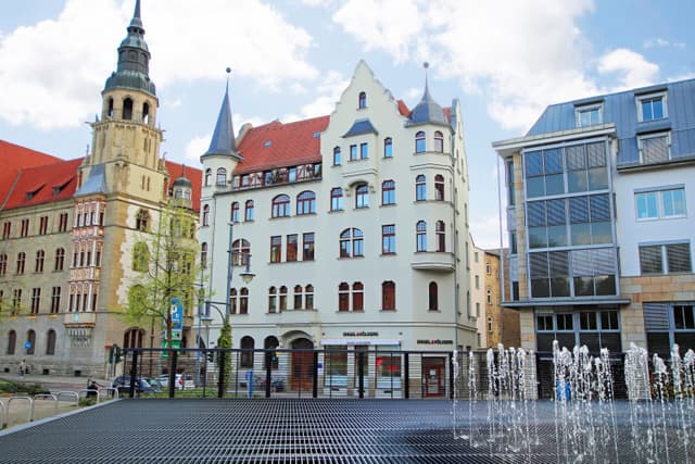 Exterior view of Engel & Völkers real estate office in a European city. The building is white with red trim and a red tile roof. A fountain is in the foreground.
