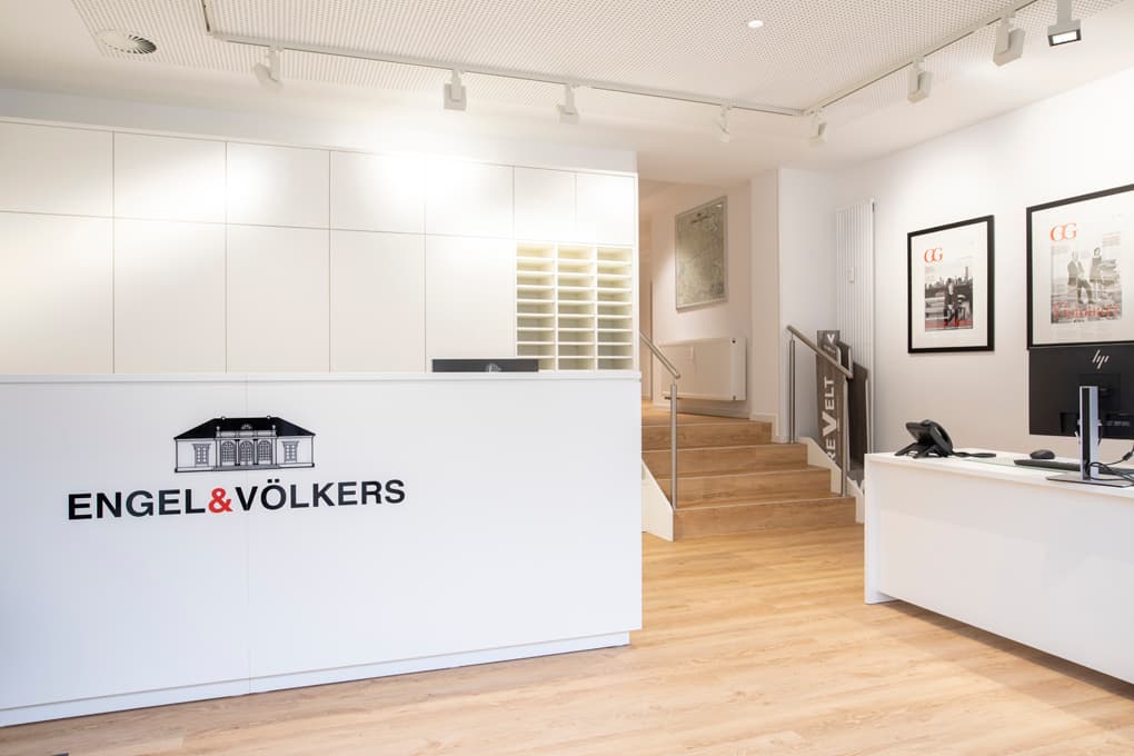 Engel & Völkers real estate office exterior. Brick building with glass storefront. White planters flank the entrance.